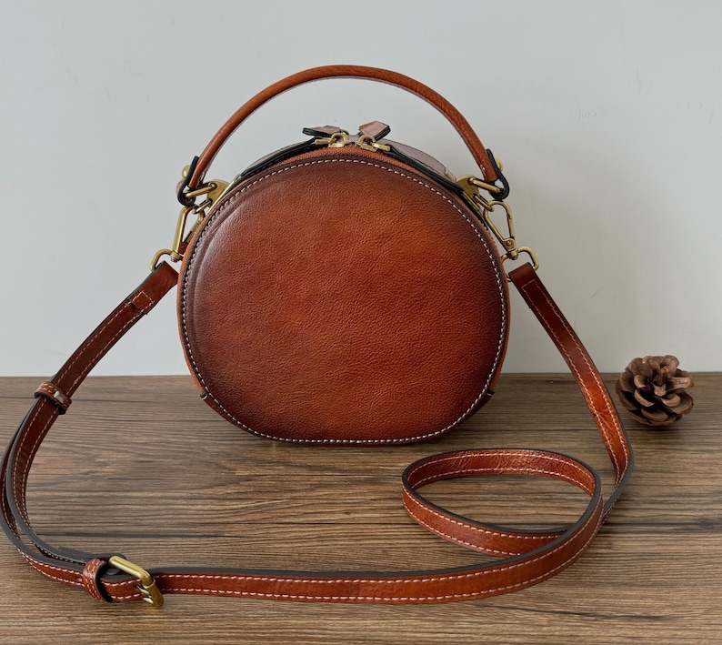 May include: A round, brown leather handbag with a top handle and a detachable shoulder strap. The bag features a zipper closure and gold-colored hardware. The stitching is white, and a small pinecone sits on the wooden surface.