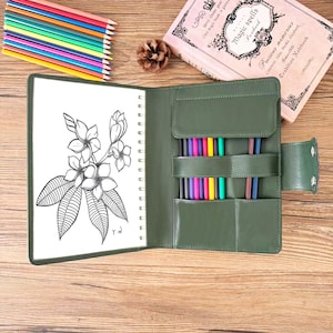 May include: An open, olive-green leather journal with a spiral-bound sketchbook featuring a floral illustration. Colored pencils are neatly arranged, alongside a decorative pinecone and a vintage-style notebook with the text "Magic Spells".