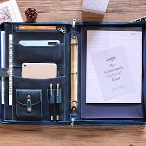 May include: A blue leather portfolio with multiple pockets, a zipper closure, and a ring binder. The portfolio contains a smartphone, tablet, pens, and a notepad with the book title "The Astonishing Color of After".