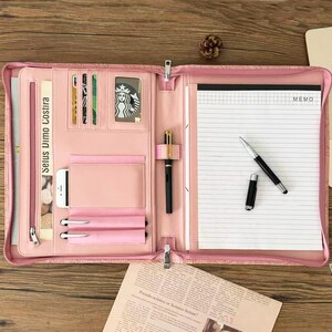 May include: A pink zippered portfolio organizer featuring multiple interior card slots, a clear ID window, a smartphone pocket, two pen loops, and a notepad with lined memo paper. A black fountain pen rests on the notepad, and a second pen is held in a loop.