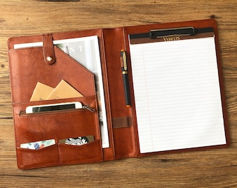 May include: A brown leather portfolio with a notepad, pen, and multiple pockets. The notepad has a clip and the text "Veritas" printed on the top.