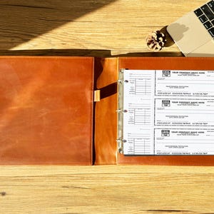 Può includere: Raccoglitore in pelle marrone aperto con meccanismo a tre anelli, contenente assegni stampati. Il raccoglitore è aperto su una superficie di legno, con un computer portatile e una pigna sullo sfondo. Gli assegni hanno campi prestampati.