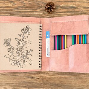 May include: Open pink leather art portfolio with a spiral-bound sketchbook featuring a floral and butterfly illustration. The portfolio includes a pocket with a variety of colored pencils. A pinecone and colored pencils are visible in the background.
