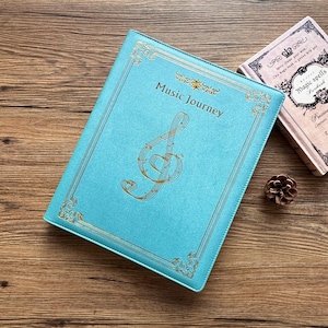 May include: A turquoise journal with the words "Music Journey" and a gold treble clef design. The journal has gold decorative borders and is next to a small book with the words "Magic spells". A pine cone rests nearby.