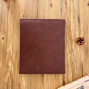 Puede incluir: Un cuaderno o portafolio rectangular de cuero marrón oscuro. La cubierta es lisa y parece estar cosida a lo largo de los bordes. El fondo es una superficie de madera con una piña.
