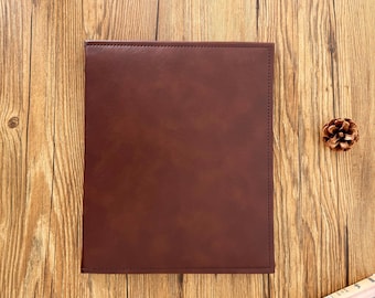 Könnte beinhalten: Ein rechteckiges, dunkelbraunes, ledergebundenes Notizbuch oder Portfolio. Der Einband ist glatt und scheint an den Rändern genäht zu sein. Der Hintergrund ist eine Holzoberfläche mit einem Tannenzapfen.