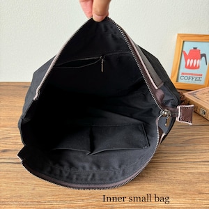 May include: A black canvas bag with a brown leather trim and zipper closure. The bag has a zippered pocket on the inside and a smaller pocket on the side. The bag is open and the inside is visible.