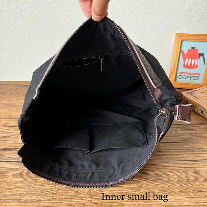 May include: A black canvas bag with a brown leather trim and zipper closure. The bag has a zippered pocket on the inside and a smaller pocket on the side. The bag is open and the inside is visible.