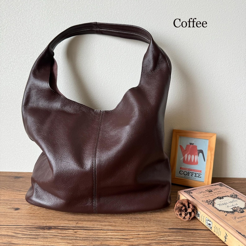 May include: A brown leather hobo bag with a single strap. The bag is sitting on a wooden surface with a book and a small wooden decoration.