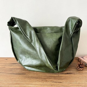 Puede incluir: Un bolso de hombro verde con un asa grande y curvada. El bolso está hecho de un material suave y texturizado y tiene un diseño holgado. El bolso está sobre una superficie de madera.
