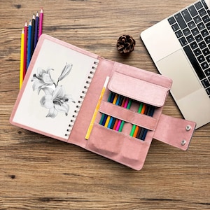 May include: Open pink leather art journal with a spiral binding, featuring a pencil sketch of lilies. The journal has a built-in pencil holder filled with colored pencils. A laptop and pinecone are also visible.