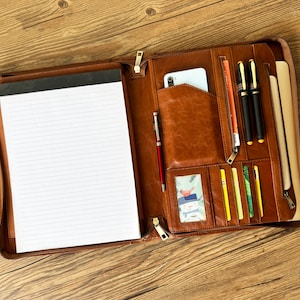May include: Open brown leather portfolio with a zippered closure. Inside, there's a notepad, pen holders, a smartphone pocket, and slots for cards. The portfolio also holds pens and a small picture. The background is a wooden surface.