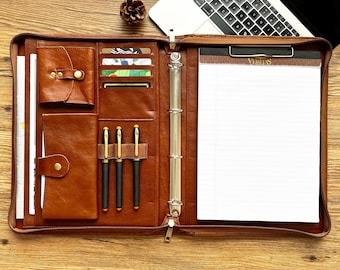 May include: A brown leather portfolio with a zipper closure, a ring binder, and a notepad. The notepad has the word "Veritas" printed on it. The portfolio has multiple pockets and slots for pens, cards, and other items.