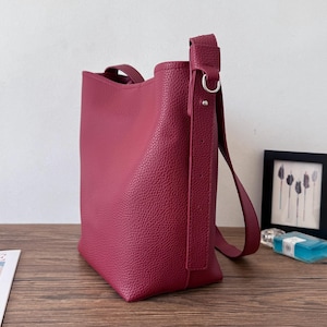 Puede incluir: Bolso tote de color burdeos con superficie texturizada. El bolso tiene una correa para el hombro y un asa superior. Se encuentra sobre una superficie de madera.