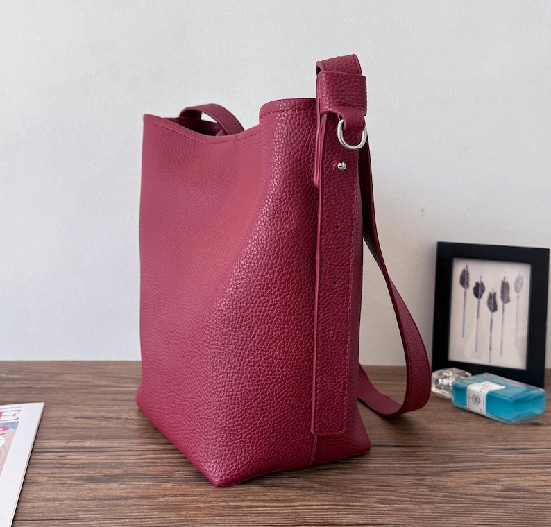 Puede incluir: Bolso tote de color burdeos con superficie texturizada. El bolso tiene una correa para el hombro y un asa superior. Se encuentra sobre una superficie de madera.