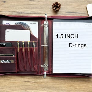 Puede incluir: Un portafolio de cuero burdeos con cierre de cremallera, con múltiples bolsillos para una tableta, teléfono, bolígrafos y tarjetas. Incluye un bloc de notas con anillas en D de 3,8 cm y la palabra "MEMO".