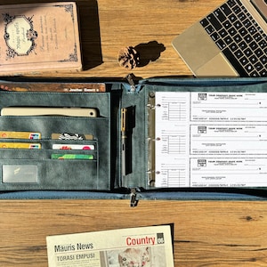 May include: A dark teal leather binder, open to reveal check forms and card pockets. A pen is clipped inside. A laptop and a book are visible. The binder is on a wooden surface.
