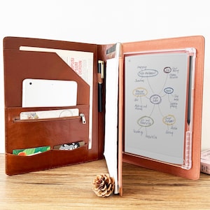 May include: A brown leather portfolio organizer featuring a tablet, a zippered pocket, card slots, a pen loop with a black pen, a notepad, and a digital e-ink tablet displaying a handwritten mind map diagram with various text labels.