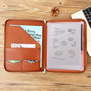 May include: Open brown leather portfolio with a zipper, containing a digital tablet with a stylus and a book titled "Boats Things that Float". The portfolio has pockets for cards and documents. A laptop is visible in the background.