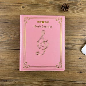 May include: Pink notebook with gold trim and a gold treble clef design. The cover reads "Music Journey" in gold lettering. The notebook is rectangular and sits on a wooden surface.