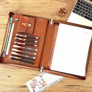 May include: A tan leather portfolio case featuring a small flap pocket, multiple elastic pen loops holding various writing instruments, and a lined memo pad with the word MEMO printed at the top, all secured by a zipper closure.