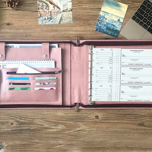 May include: A pink leather binder with a zipper closure, featuring multiple pockets and slots for holding cards, pens, and a notepad. The binder is open to reveal a chequebook and a calendar.