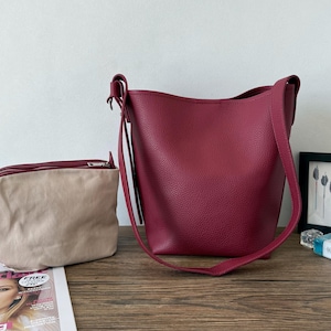 Puede incluir: Un bolso tipo cubo de cuero burdeos con una correa larga para el hombro y una bolsa beige m&aacute;s peque&ntilde;a con cremallera. Los bolsos est&aacute;n sobre una superficie de madera, con una revista y una imagen enmarcada en el fondo.