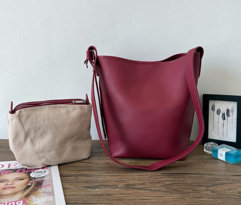 Puede incluir: Un bolso tipo cubo de cuero burdeos con una correa larga para el hombro y una bolsa beige m&aacute;s peque&ntilde;a con cremallera. Los bolsos est&aacute;n sobre una superficie de madera, con una revista y una imagen enmarcada en el fondo.