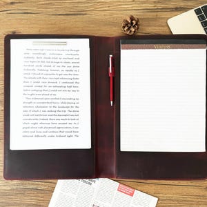 Puede incluir: Un portafolio de cuero burdeos con un bloc de notas, un bolígrafo y un documento. El bloc de notas tiene la palabra "VERITAS" impresa. Un bolígrafo rojo está insertado en el portafolio. También se ven un portátil y una piña.