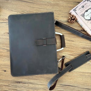 May include: A dark brown leather briefcase with a handle and a shoulder strap. The briefcase has a flap closure with a leather strap and a metal clasp. The stitching is white. A book and pinecone are in the background.