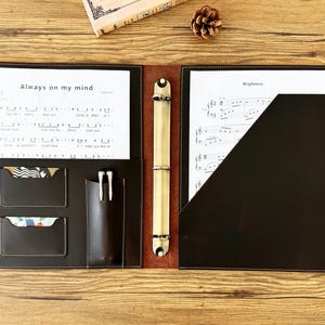 Peut inclure: Un portfolio en cuir marron ouvert avec des partitions et un porte-stylo. Le côté gauche comporte deux poches pour cartes et un porte-stylo avec deux stylos. Le côté droit a une poche. Le texte "Always on my mind" est visible.
