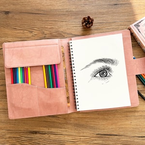 Puede incluir: Portafolio de arte rosa abierto con un cuaderno de bocetos encuadernado en espiral con un dibujo de un ojo. El portafolio tiene un bolsillo lleno de lápices de colores. Una piña descansa sobre la superficie de madera.