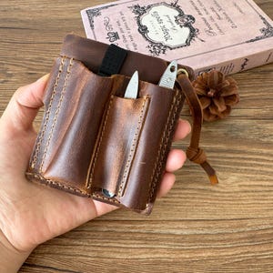 May include: A brown leather pocket organizer featuring two vertical slots with visible stitching, a black elastic loop at the top, and a leather pull tab. Two small metal tools are inserted into the slots, showcasing a rugged, textured finish.