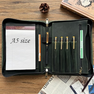 May include: A dark green leather A5 size notebook with a zipper closure. The notebook has a built-in pen holder and several slots for pens and pencils. The notebook is open and the inside is lined with white paper. The text "VERITAS" and "A5 size" is visible on the notebook.