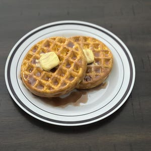 May include: Two golden-brown waffles on a white plate with a black rim. Each waffle is topped with a pat of butter and drizzled with syrup. The plate sits on a dark wooden surface.
