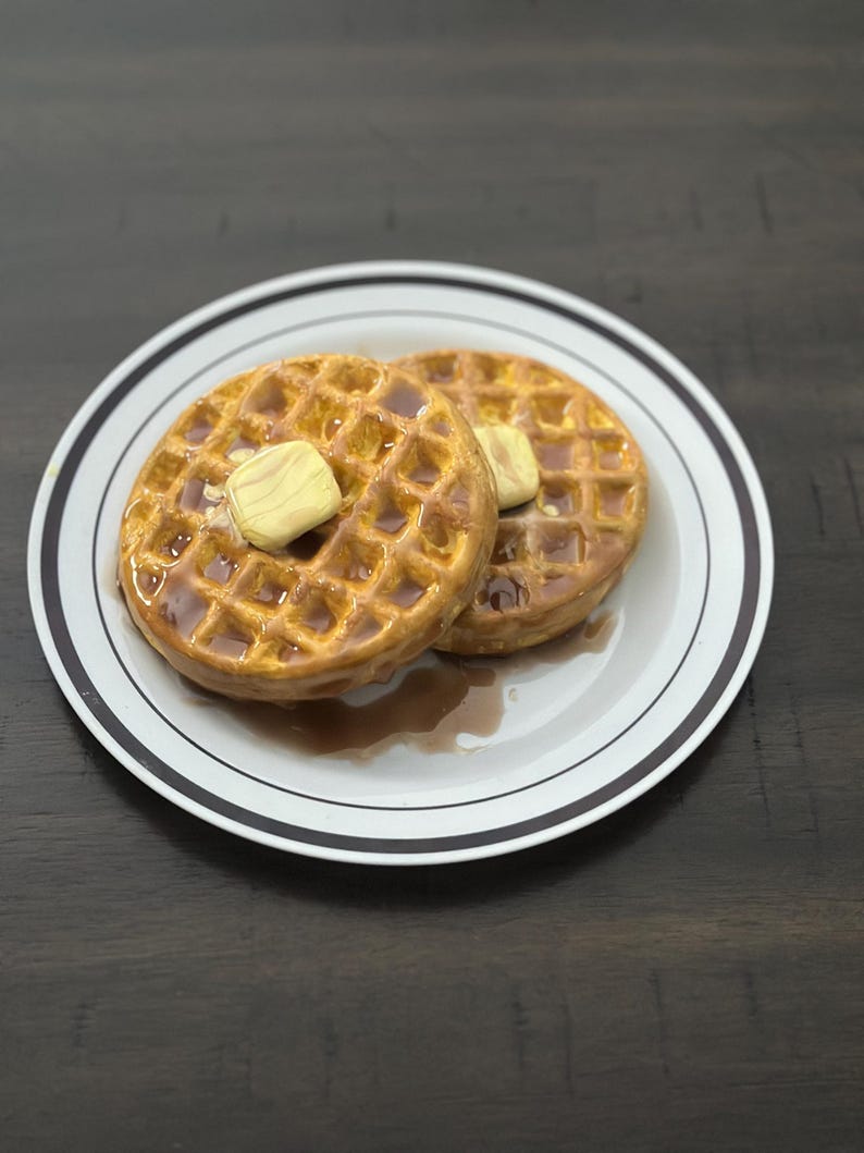 May include: Two golden-brown waffles on a white plate with a black rim. Each waffle is topped with a pat of butter and drizzled with syrup. The plate sits on a dark wooden surface.