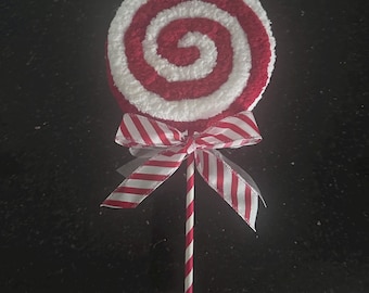 May include: A circular lollipop decoration featuring a textured red and white spiral pattern. The lollipop is attached to a thin, straight stick with a red and white diagonal striped design. A decorative ribbon with a red and white striped pattern is tied in a bow at the base of the lollipop head.