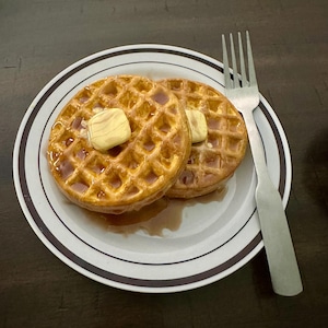 May include: Two golden-brown waffles topped with butter and syrup on a white plate with a black and brown rim. A silver fork rests beside the waffles, ready for a delicious breakfast.