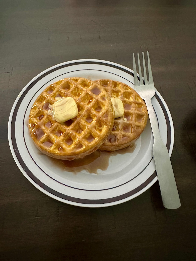 May include: Two golden-brown waffles topped with butter and syrup on a white plate with a black and brown rim. A silver fork rests beside the waffles, ready for a delicious breakfast.