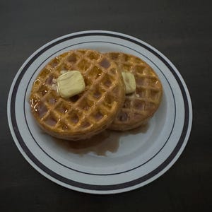 May include: Two golden-brown waffles on a white plate with a blue and black striped rim. Each waffle is topped with a pat of butter and drizzled with syrup. The plate sits on a dark surface.