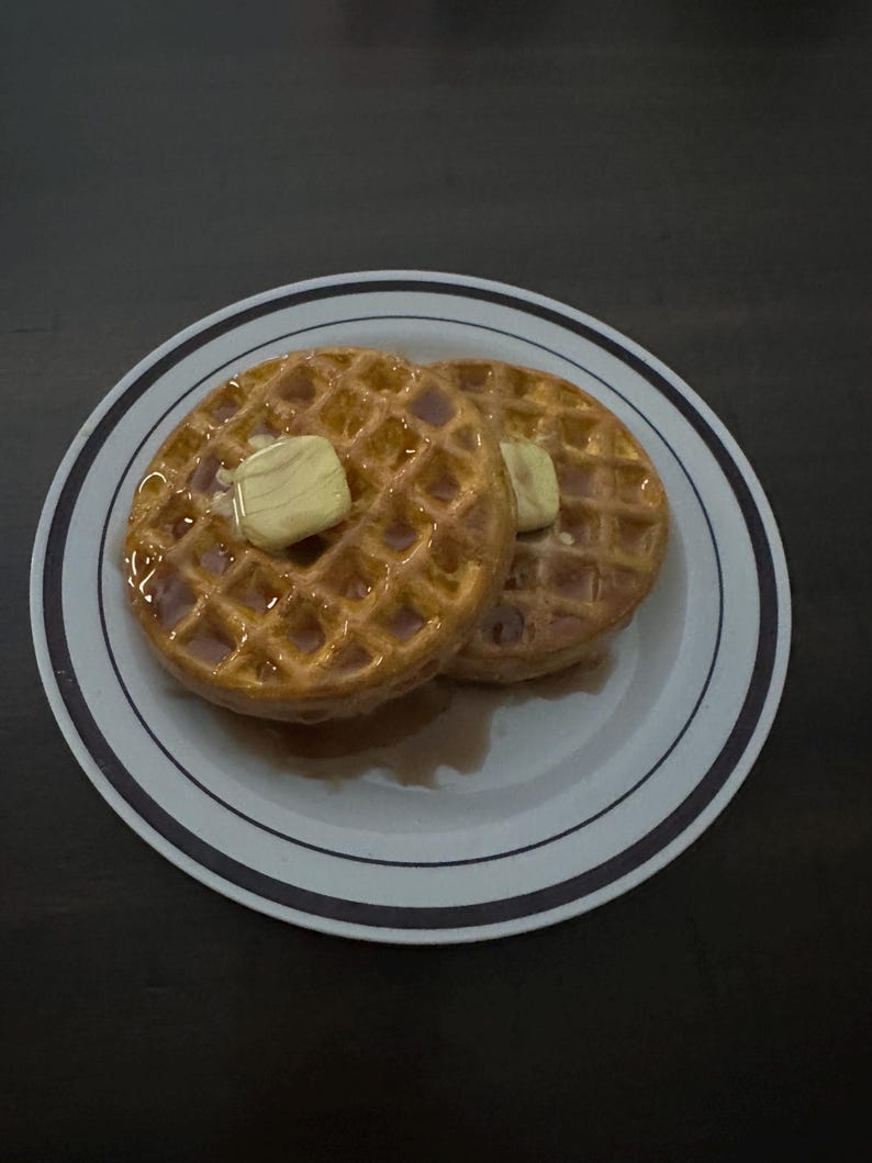 May include: Two golden-brown waffles on a white plate with a blue and black striped rim. Each waffle is topped with a pat of butter and drizzled with syrup. The plate sits on a dark surface.