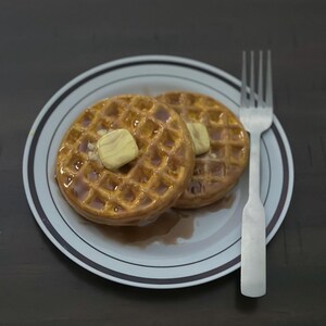 May include: Two golden-brown waffles topped with butter and syrup on a white plate with a brown and white rim. A silver fork rests beside the plate. The waffles are a breakfast food.