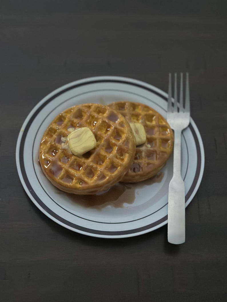 May include: Two golden-brown waffles topped with butter and syrup on a white plate with a brown and white rim. A silver fork rests beside the plate. The waffles are a breakfast food.