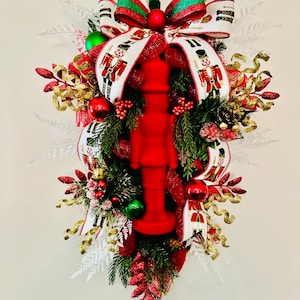 May include: A red felt nutcracker figurine with a white and green bow, surrounded by red, green, and gold Christmas decorations. The wreath is decorated with white, red, and gold ribbon.
