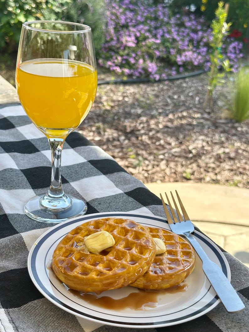 May include: A plate of two golden-brown waffles topped with butter and syrup, served with a glass of orange juice. A silver fork rests on the plate. The scene is set on a black and white checkered tablecloth.