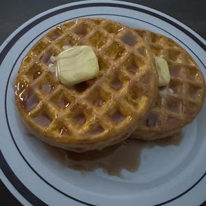 May include: Two golden-brown waffles topped with butter and syrup on a white plate with blue stripes. The waffles have a grid pattern and are glistening with syrup, suggesting a breakfast meal.