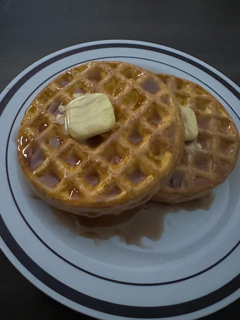 May include: Two golden-brown waffles topped with butter and syrup on a white plate with blue stripes. The waffles have a grid pattern and are glistening with syrup, suggesting a breakfast meal.