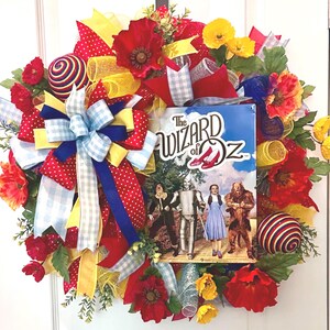 May include: A decorative wreath featuring a central sign with The Wizard of Oz text and imagery, surrounded by red poppies, yellow flowers, multi-colored striped spheres, and a large bow made of blue and white gingham, red polka dot, and yellow ribbons.