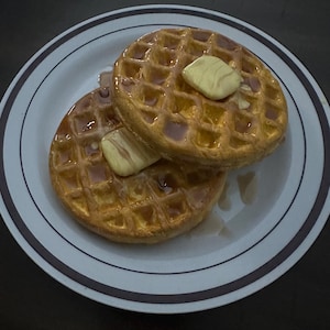 May include: Two golden-brown waffles on a white plate with a brown rim. Each waffle is topped with a pat of butter and drizzled with syrup. The waffles are a breakfast food.