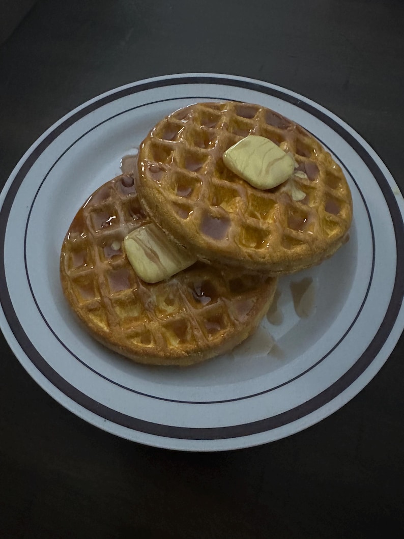 May include: Two golden-brown waffles on a white plate with a brown rim. Each waffle is topped with a pat of butter and drizzled with syrup. The waffles are a breakfast food.